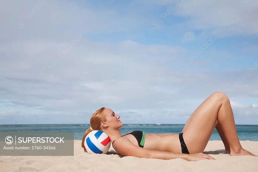 A girl in a two piece bathing suit laying on the sand resting her head on a volleyball; Waikiki, Hawaii, United States of America