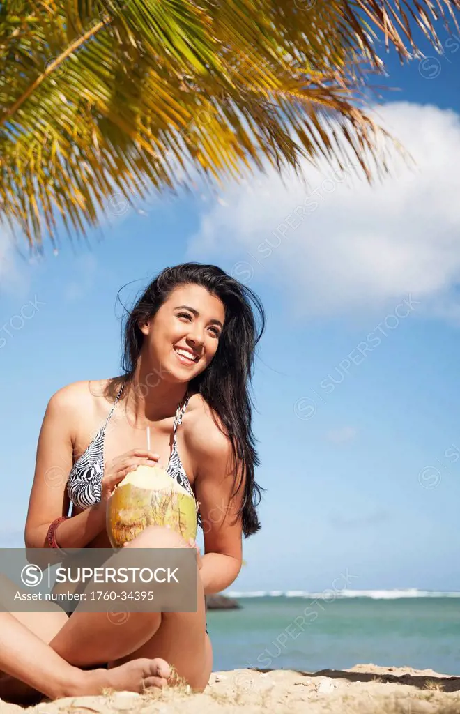 Portrait of a young woman in a bikini on the beach with a fresh coconut to drink; Oahu, Hawaii, United States of America
