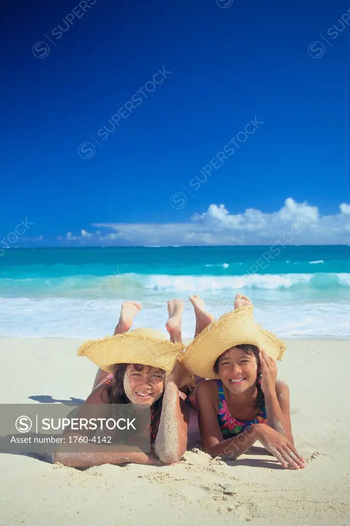 Two smiling local girls w/ floppy hats lay on beach, turquoise water & blue C1031 sky