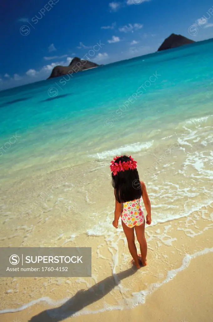 Hawaii, Oahu, Little girl with plumeria haku standing in shoreline water, Mokulua Islands in background