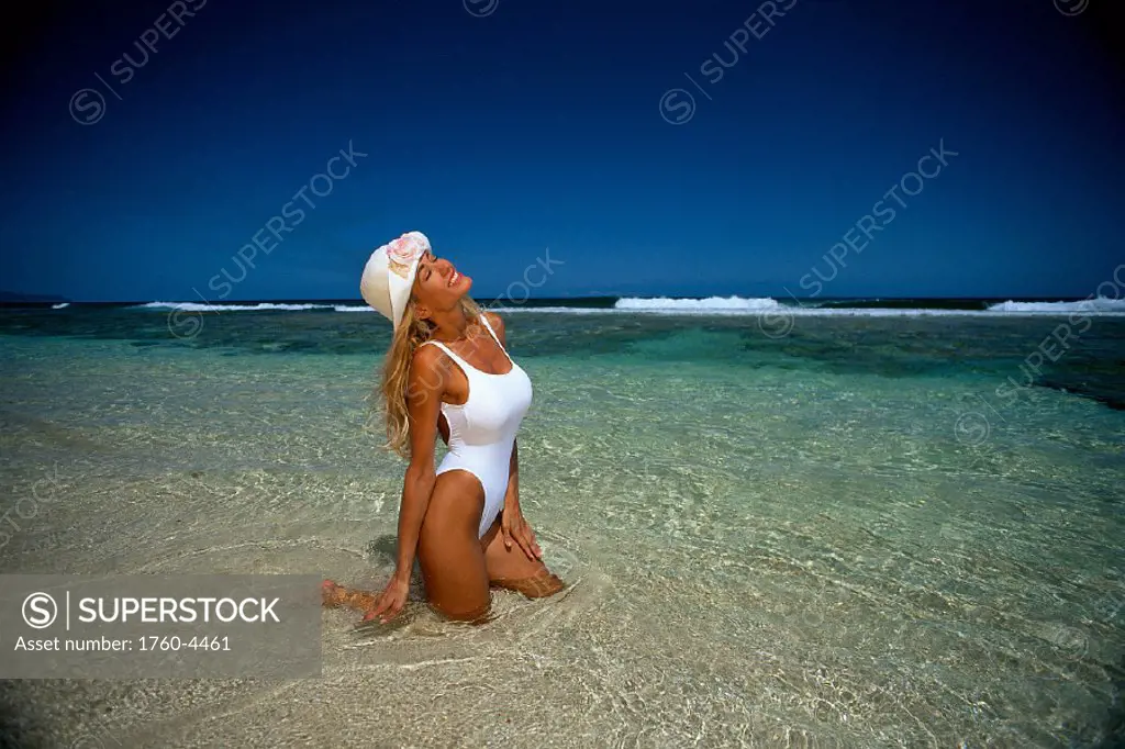 Woman in water at beach, blonde with hat kneels in shallow ocean A02A  shoreline