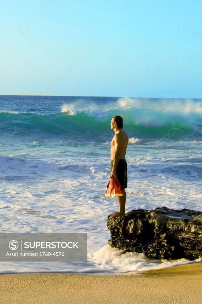 Hawaii, Oahu, Sandy Beach, Bodysurfer stands on rocks looking at ocean, holding fins.