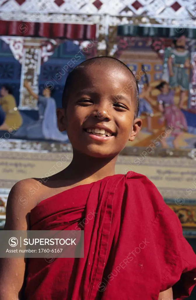 Burma (Myanmar), Bago, Shwethalyaung Paya, portrait of smiling young monk in temple, painted reliefs in background.