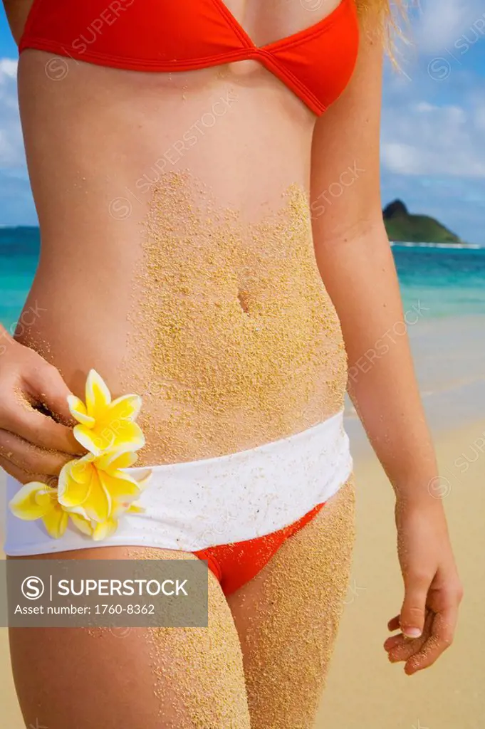 Hawaii, Oahu, Lanikai, Bikini_clad girl on beach holding plumeria flowers, Close_up on midriff covered in sand.