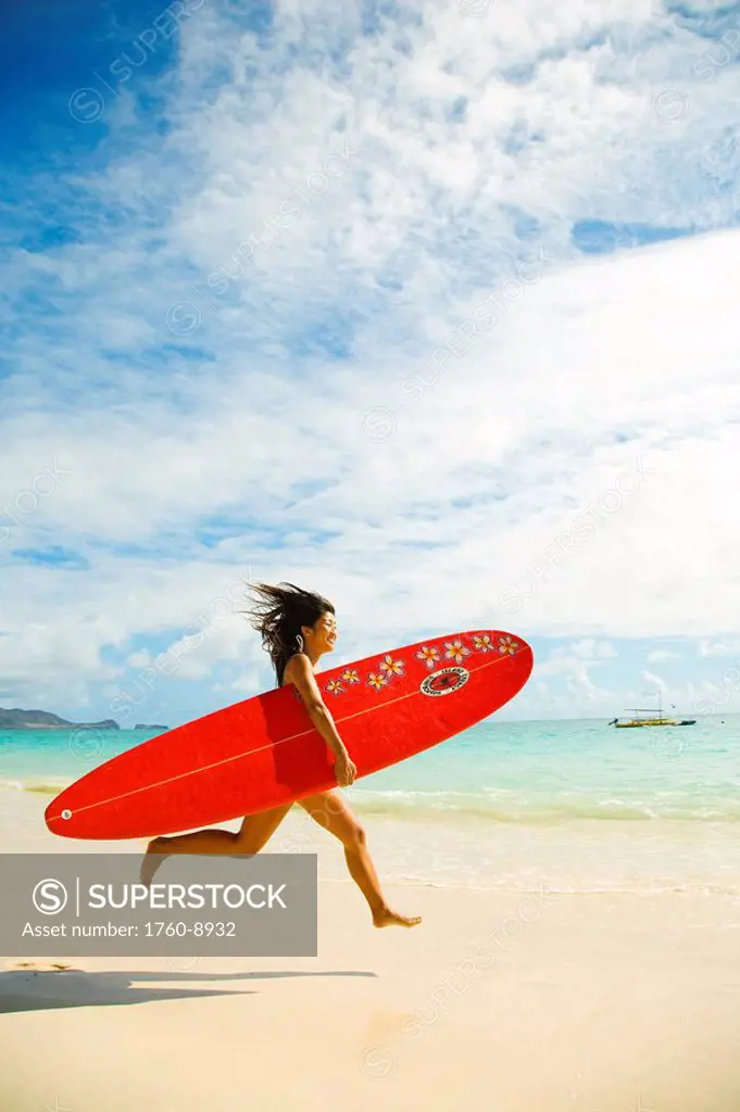 Hawaii, Oahu, Lanikai, Young Japanese woman running on beach while holding a red surfboard.