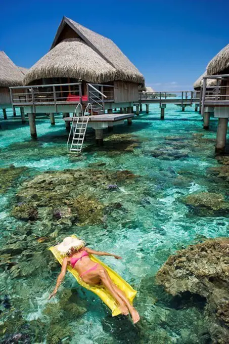 French Polynesia, Moorea, Woman sunbathing on inflatable raft over ocean reef, Luxury resort bungalows over ocean.