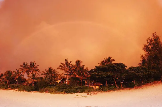 Hawaii, Oahu, North Shore, rainbow arching over vegetation and sandy beach at sunset, orange colored sky.