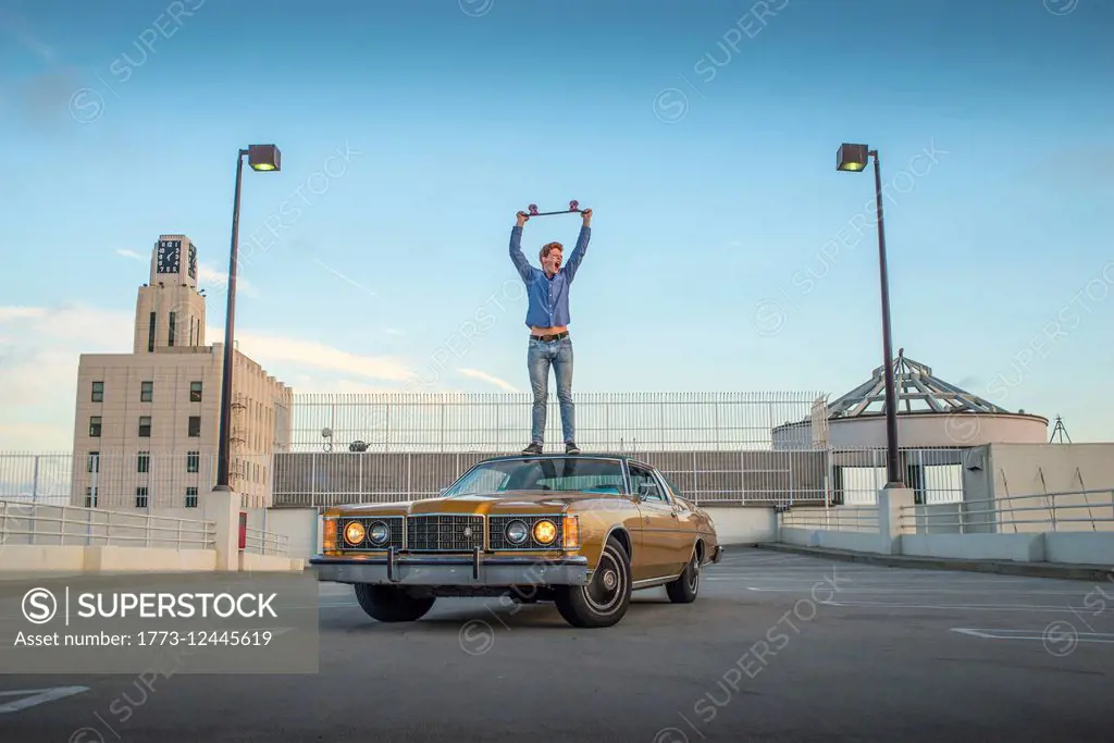 Portrait of young man standing on car holding skateboard above head