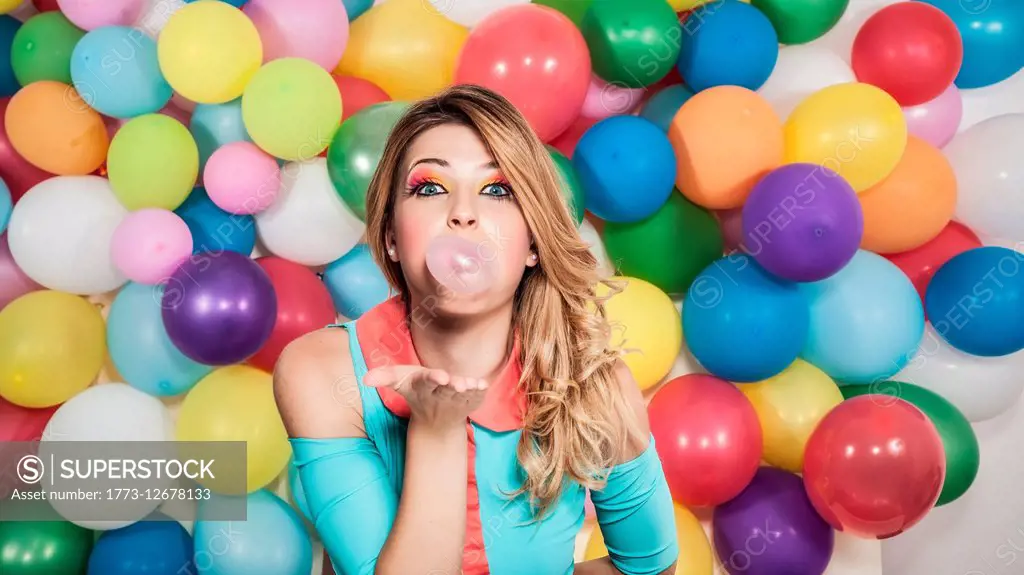 Young woman standing in front of colourful balloons, blowing bubble with bubblegum
