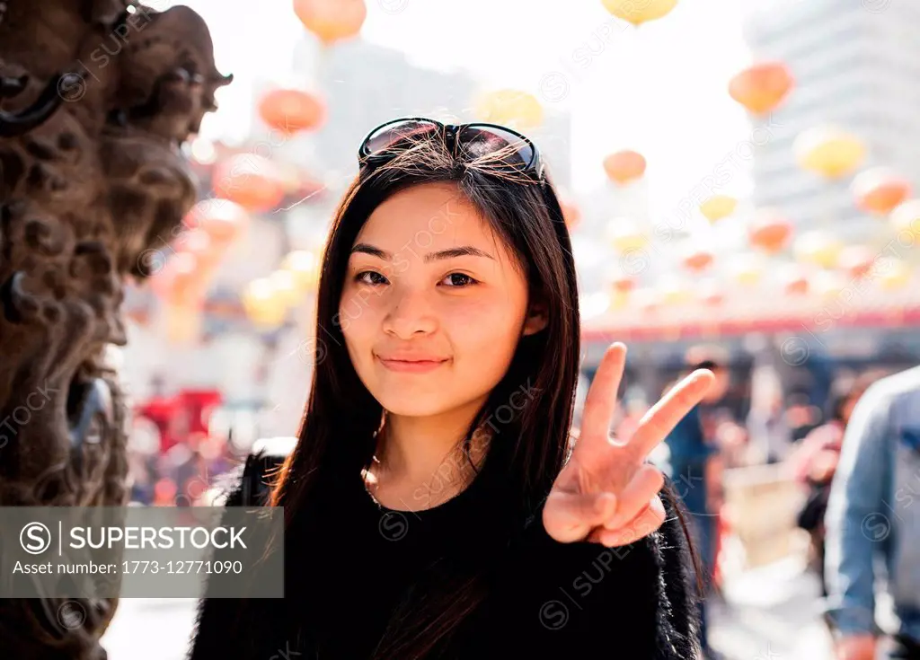 Portrait of young woman with long hair and sunglasses on head doing peace sign, looking at camera