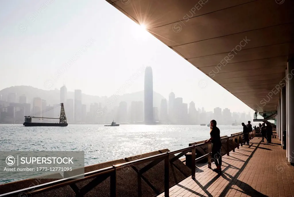 Silhouetted side view of young woman standing looking out over water at skyline, Hong Kong, China