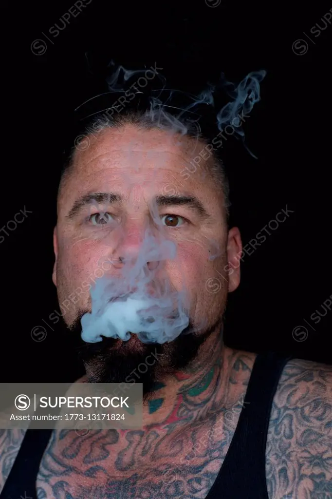 Portrait of bearded tattooed mature man looking at camera blowing smoke