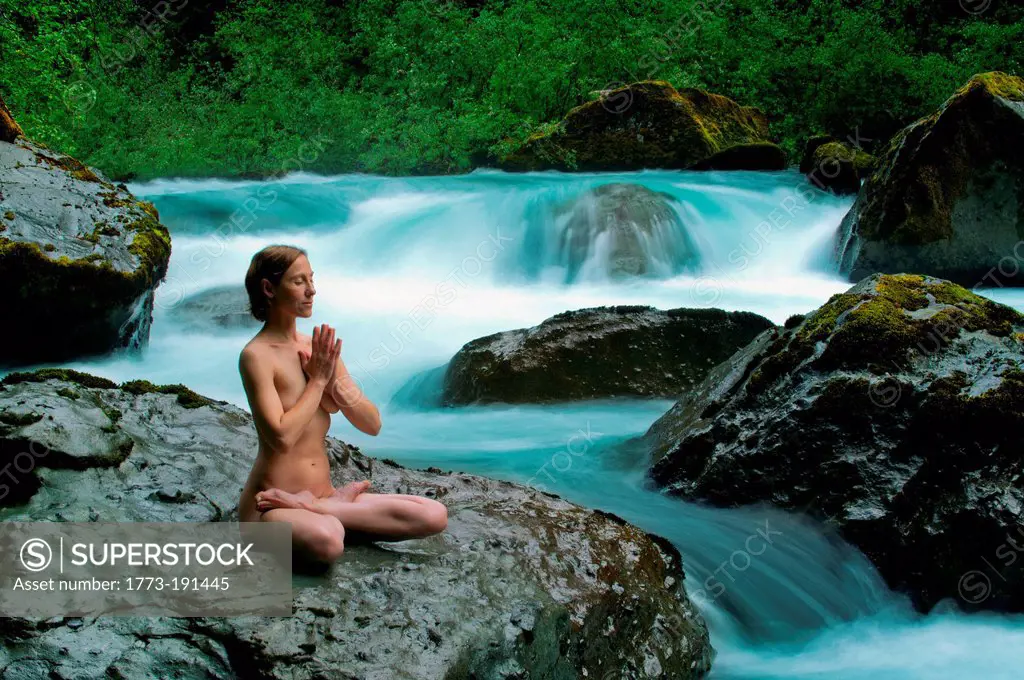 Nude woman meditating on rock by water