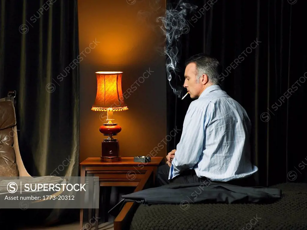 Man sitting on bed with back to camera, looking disappointed and smoking a cigarette.