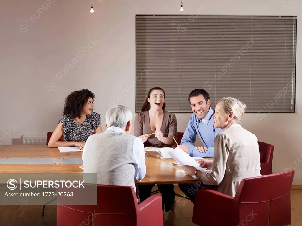 Group of people sitting around conference table having a meeting. They are all laughing and listening to an senior woman talking.