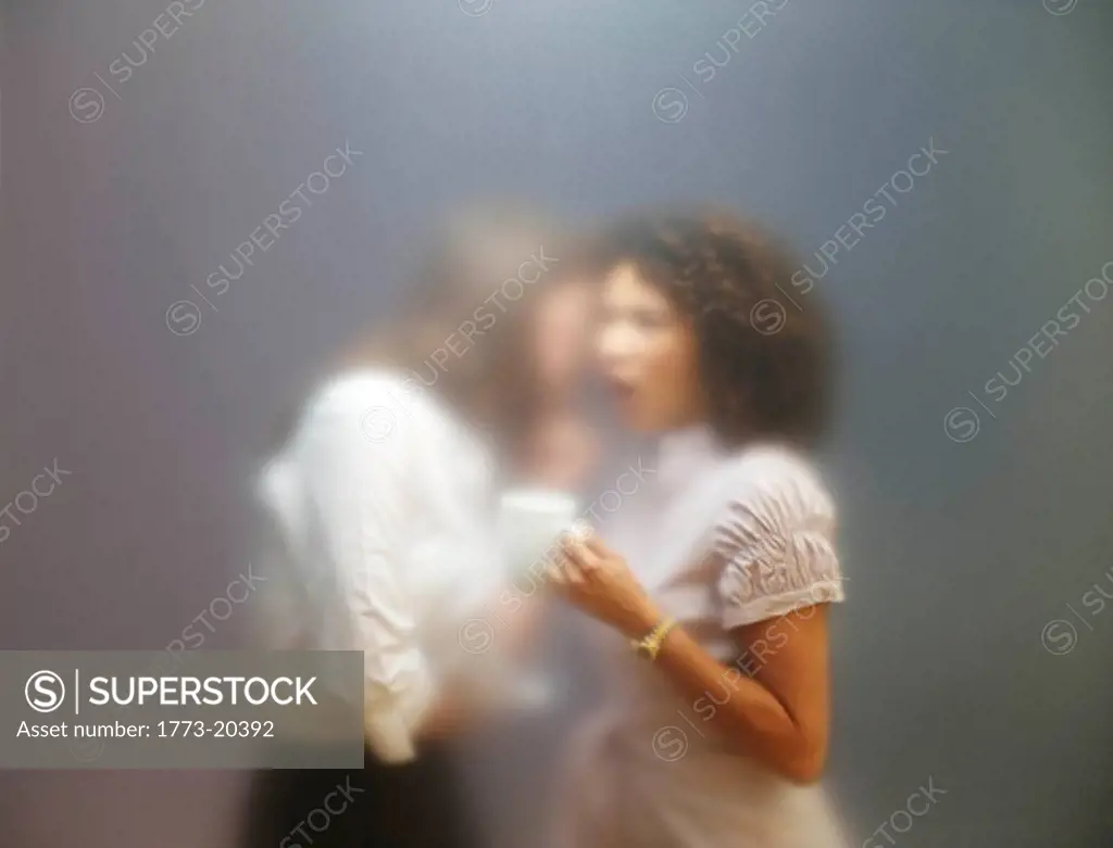 blurred image of two women behind office screen. One of them is whispering something to the other one. Shot through frosted office screen.