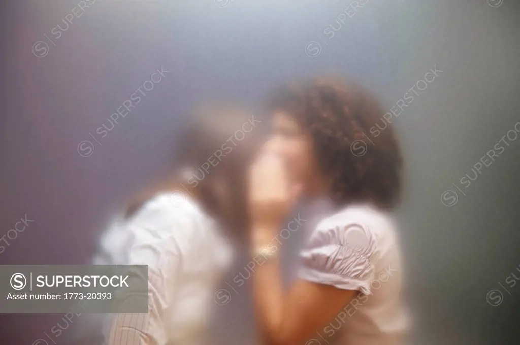 Blurred image of two women, one is whispering something to the other. Shot through frosted office screen.