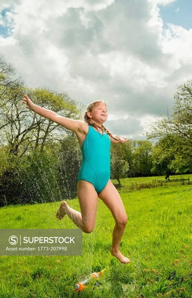 Young girl jumping over garden sprinkler in field