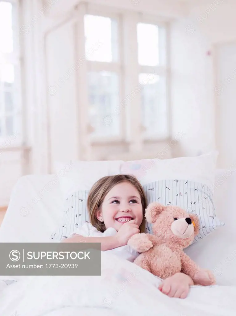 A young girl with brown hair lying in bed, a teddy bear in her arms, smiling