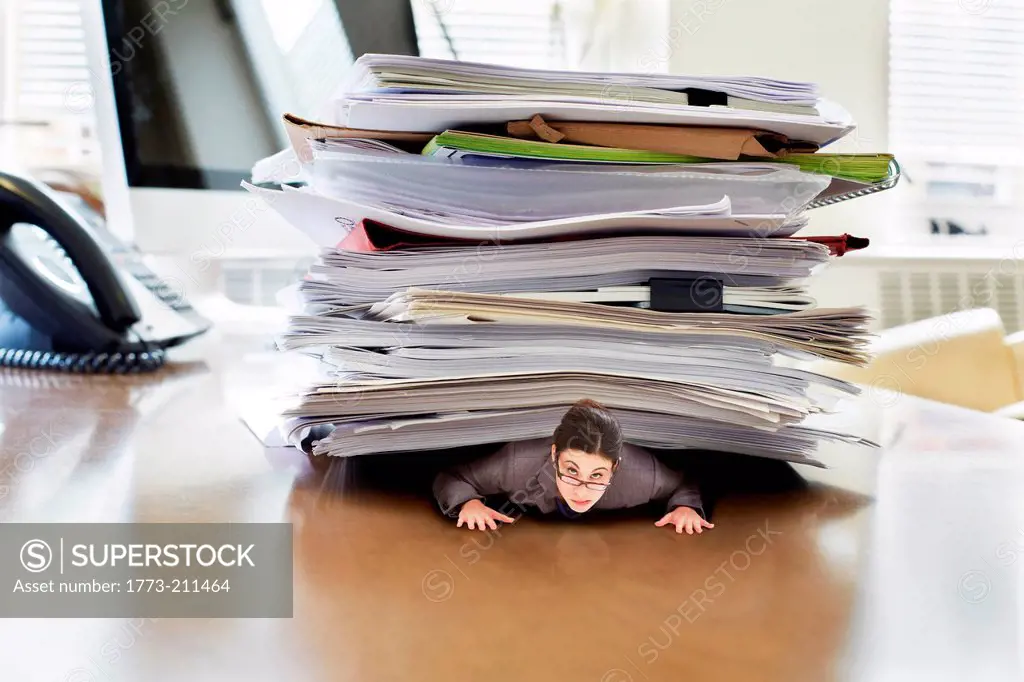 Businesswoman trapped underneath stack of large documents on oversized desk, portrait