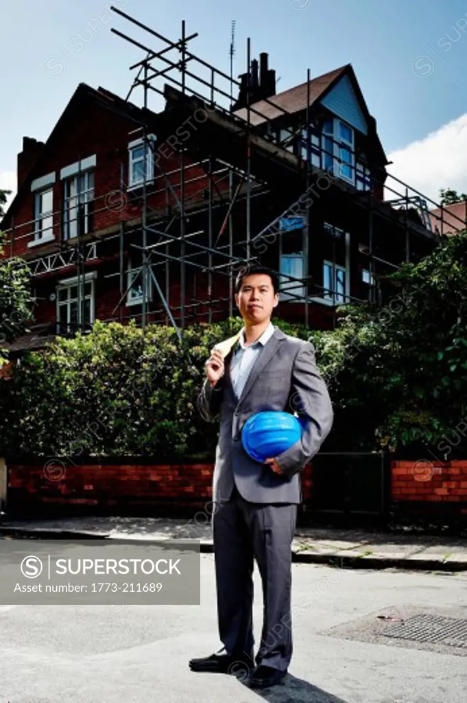 Man wearing suit, holding hard hat, standing outside house with scaffolding