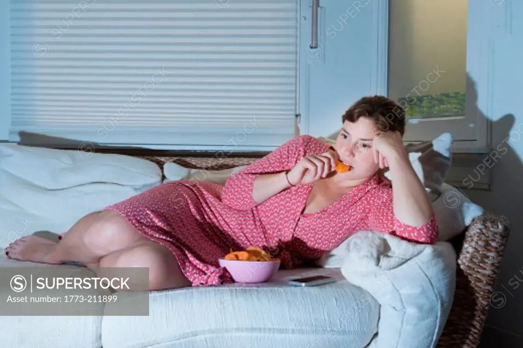 Woman lying on sofa eating bowl of crisps