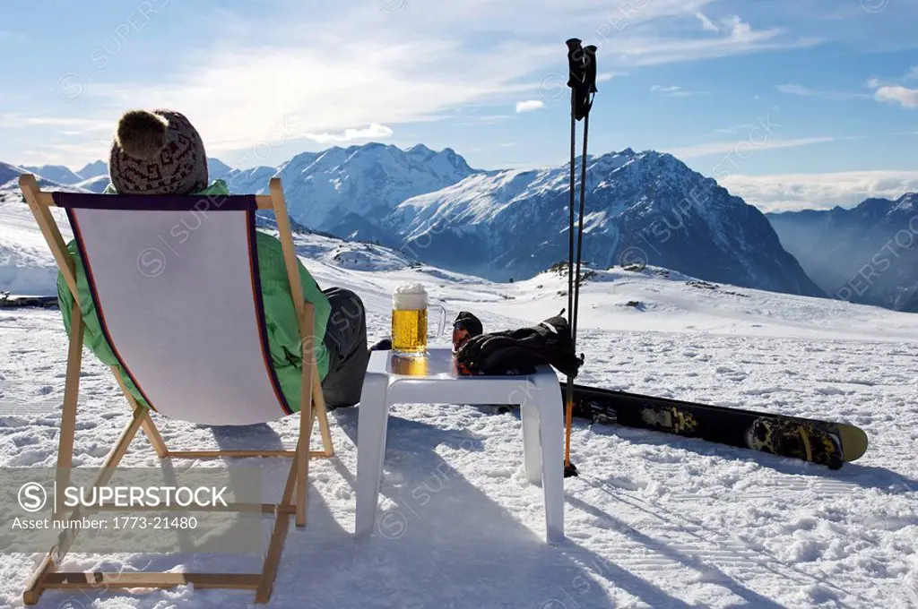Skier relaxing with a beer on a deck chair on piste.