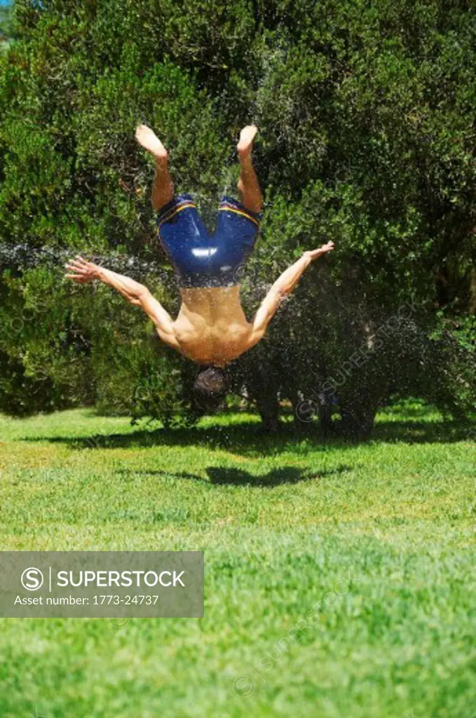 Young man doing somersault on green lawn, while being sprayed with water. He is suspended in mid air.