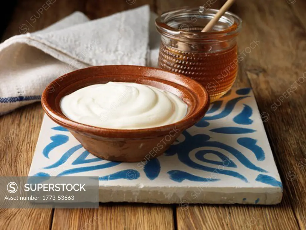 Organic Greek Yoghurt in a Terracota Bowl with a jar of Greek Honey on a blue and white ceramic tile with a napkin in the background. All on a pine wooden suface