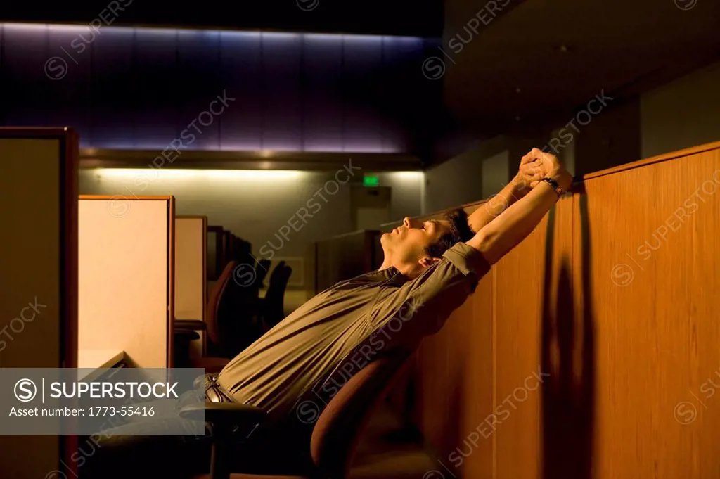 Employee taking a break when working late. Man seated at desk in row of cubicles, leaning back in chair yawning and stretching