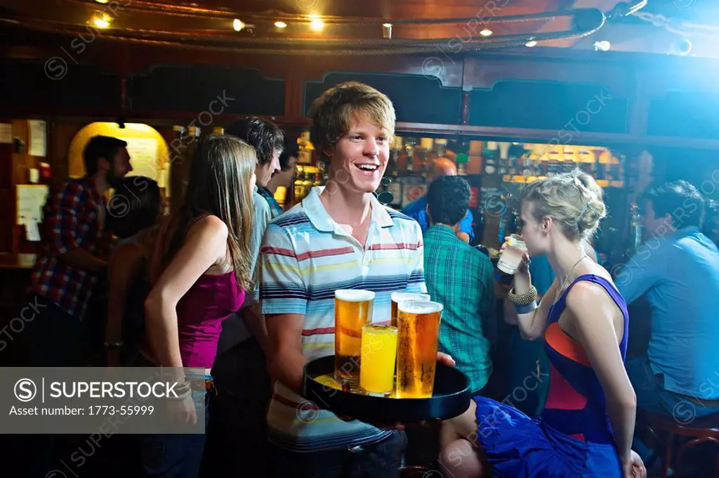 Smiling young man carrying tray with glasses of beer, he is surrounded by people. British pub interior.