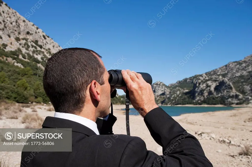 A landscape shot of a business man, dressed in a suit he appears to be spying on something far away, he is using binoculars and is standing upright on...