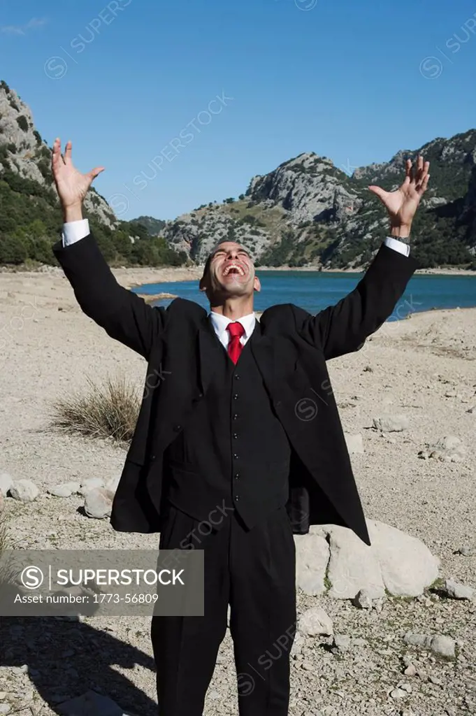 A vertical shot of a business man, dressed in a suit he appears to be punching the sky and celebrating and is standing upright on a beach. Sky is blue...