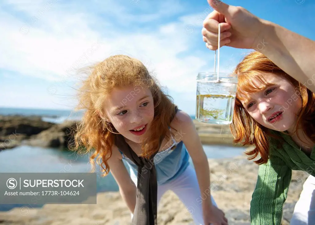 2 girls looking at fish in jar by sea