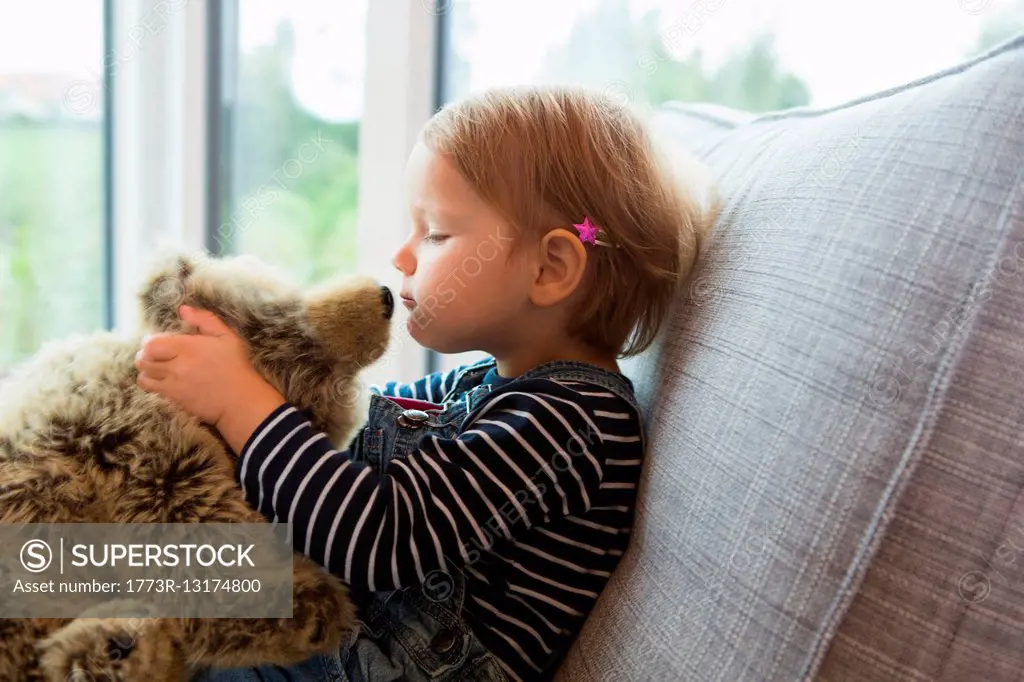 Female toddler sitting on sofa kissing teddy bear