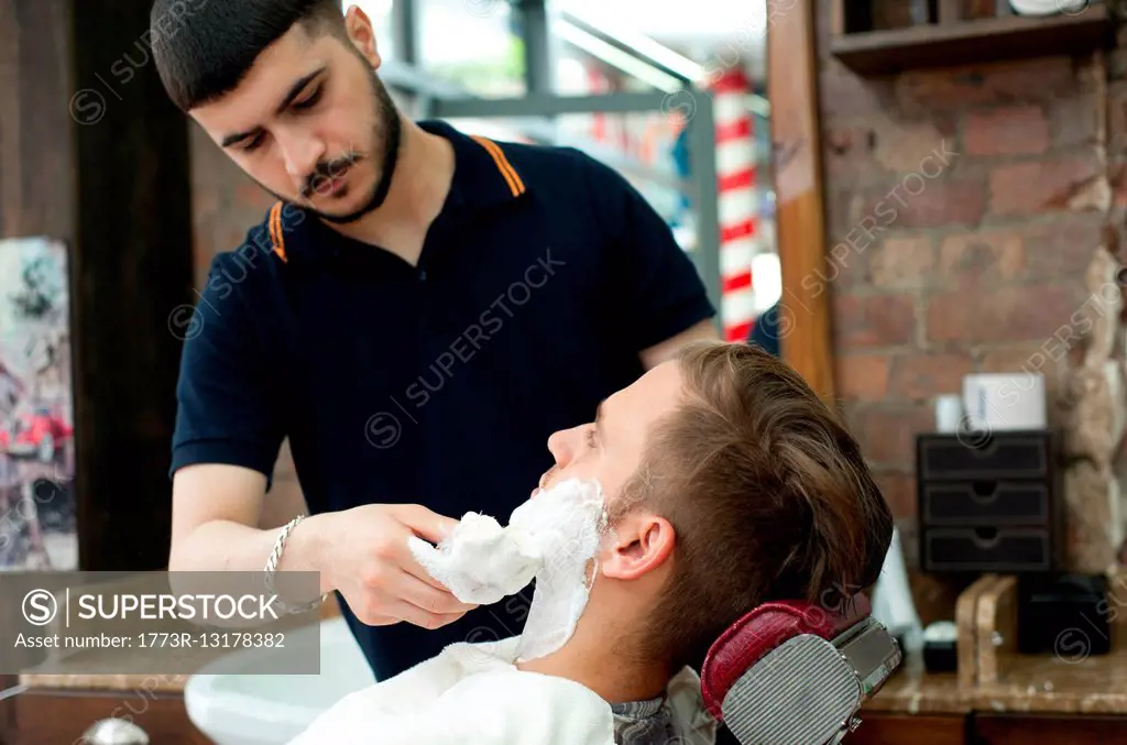 Young man in barbershop applying shaving cream to customers face