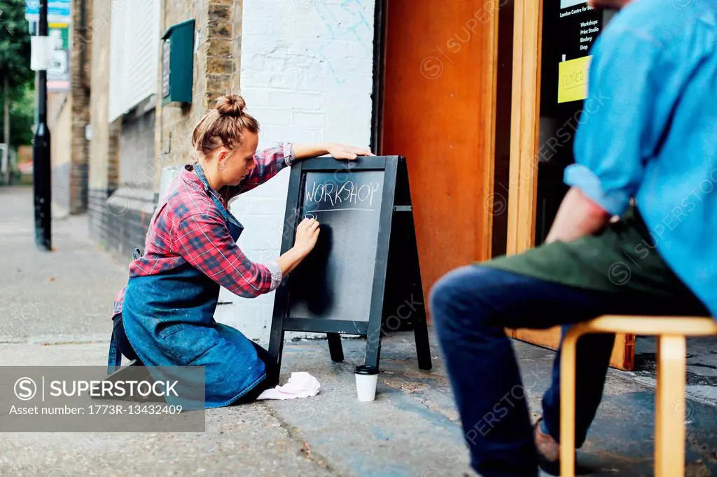 Young woman kneeling on pavement and writing on blackboard outside workshop