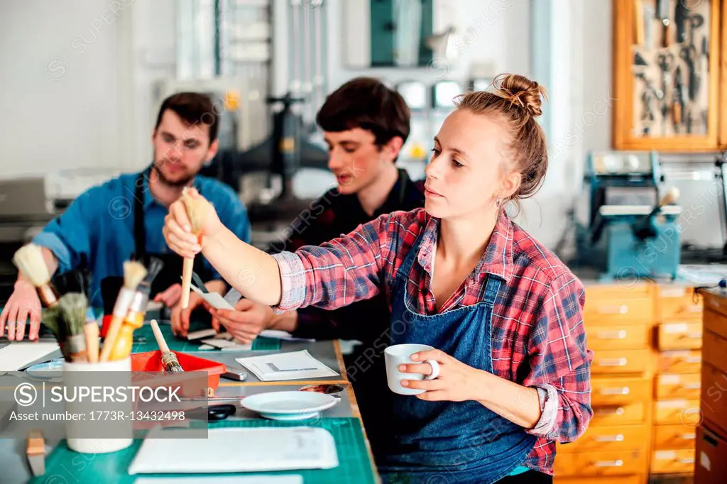 Young craftswoman holding coffee cup and reaching out for paintbrush in creative print studio, with two young craftsmen working in background