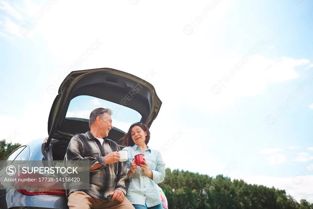 Mature couple standing beside car, holding tin cups, smiling, low angle view