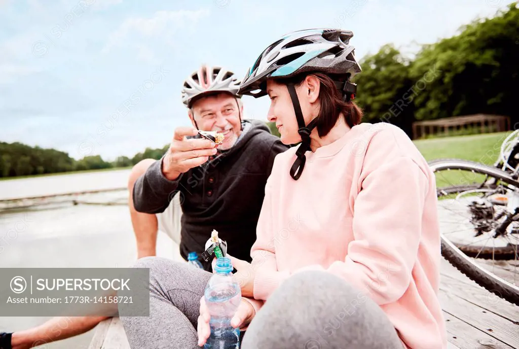 Mature couple relaxing on jetty, enjoying snack, bicycles behind them