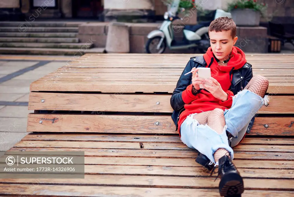 Cool young woman with short hair looking at smartphone and smoking cigarette on city bench