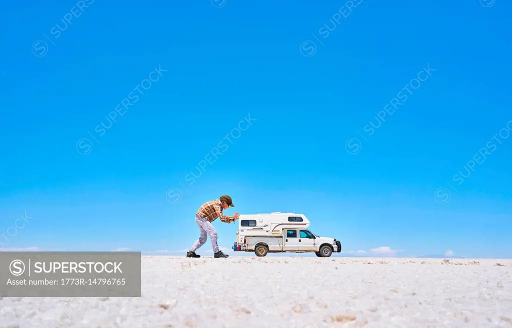 False perspective image of boy on salt flats, pretending to push recreational vehicle, vehicle in background, Salar de Uyuni, Uyuni, Oruro, Bolivia, S...