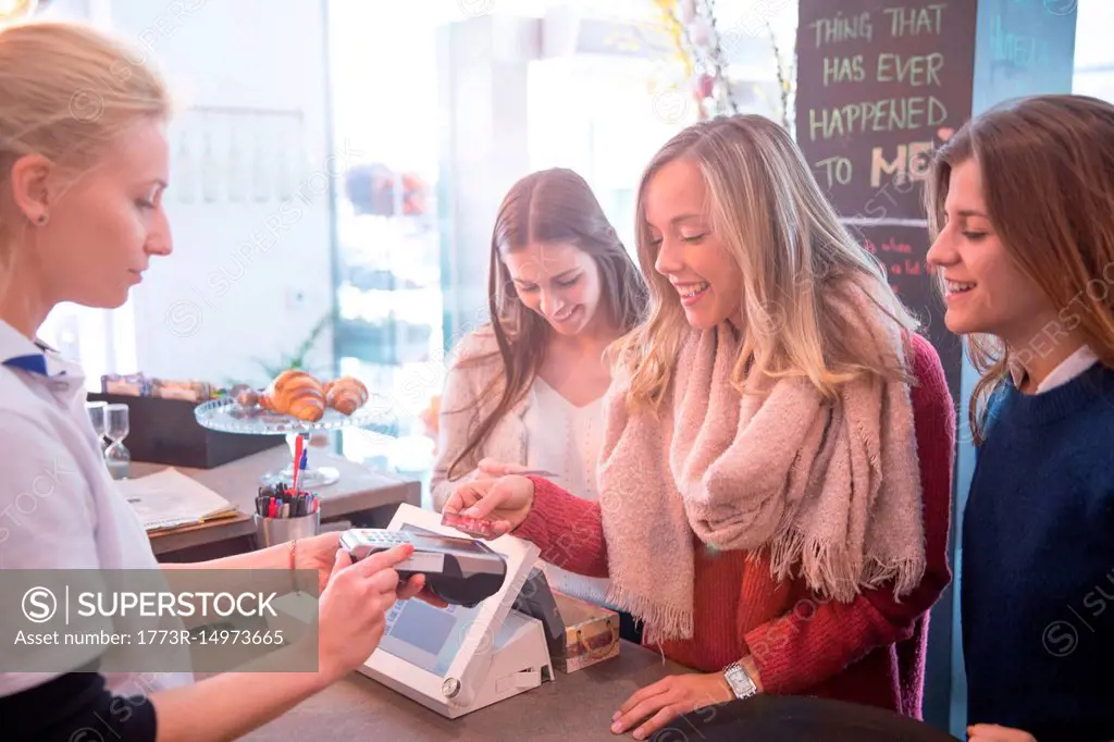Female friends standing at counter in cafe, paying using credit card