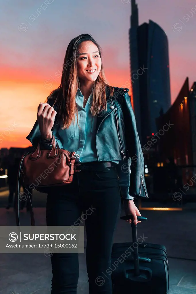 Businesswoman walking outdoors, at sunset, pulling wheeled suitcase, smiling