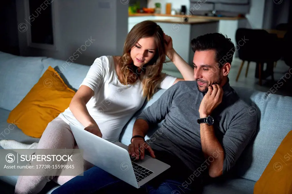 Young couple sitting on sofa in evening looking at laptop