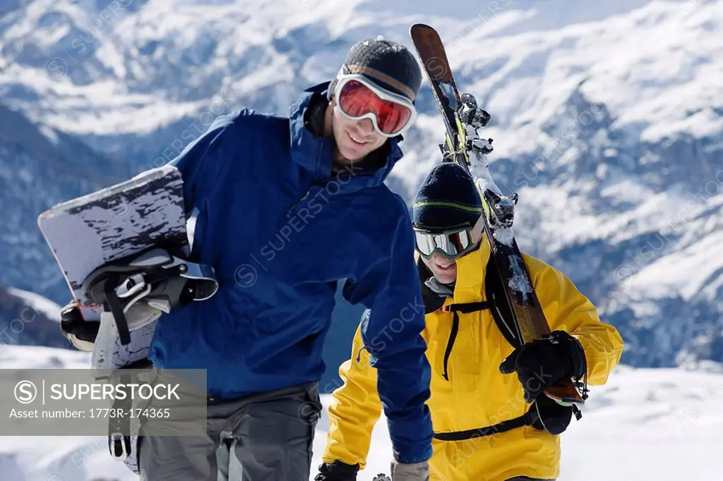 Male skier and male snowboarder walking up mountain ridge carrying skis