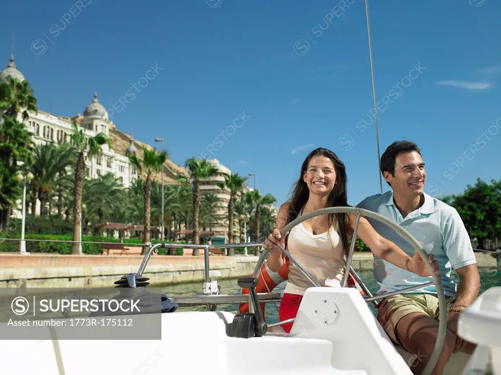 Young couple at wheel of yacht in marina, smiling