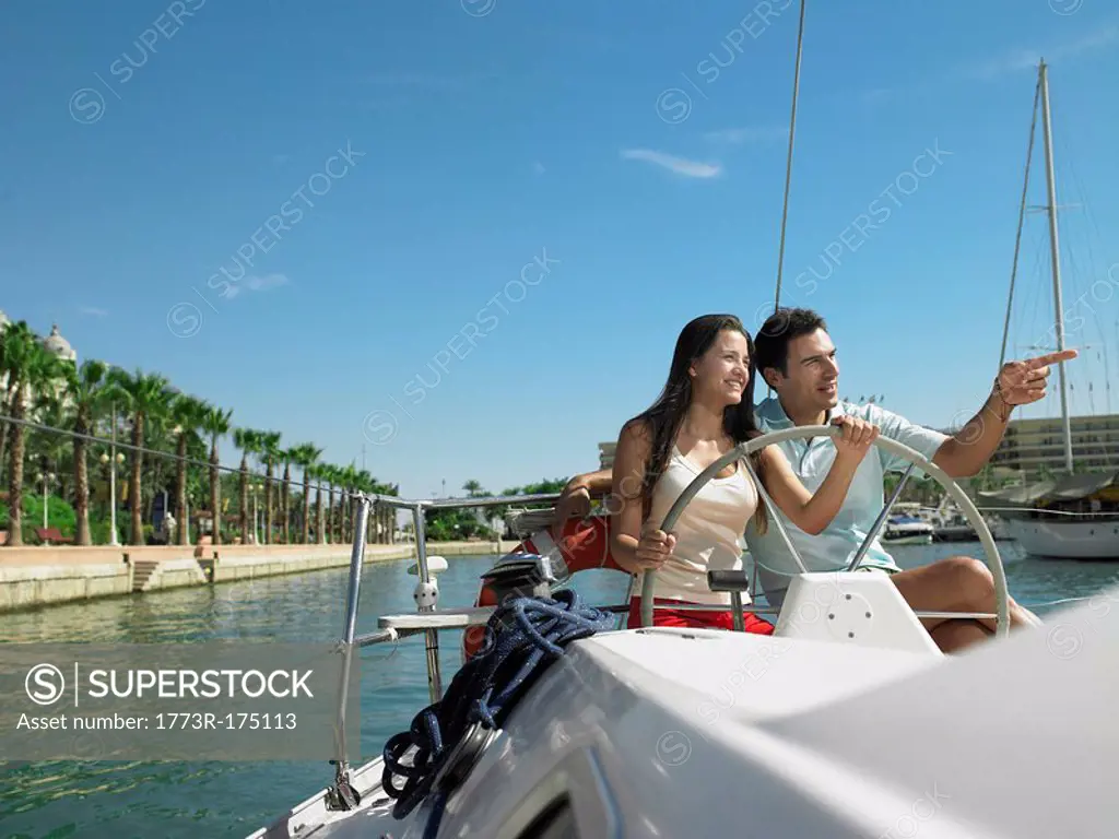 Young couple at wheel of yacht in marina, smiling