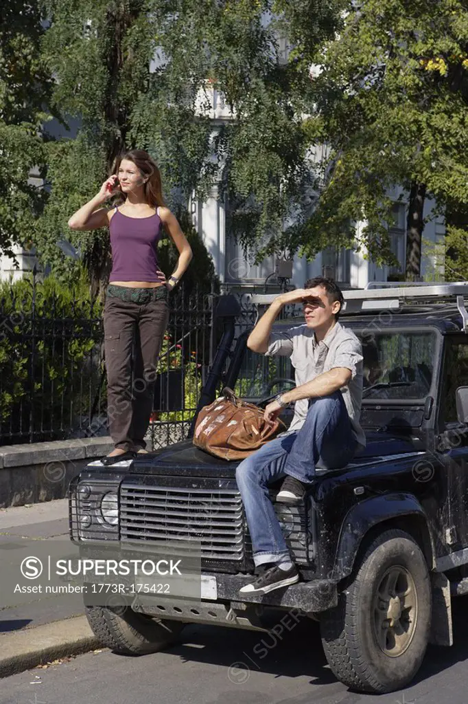 Woman standing on hood of SUV on cellular phone with man sitting on hood