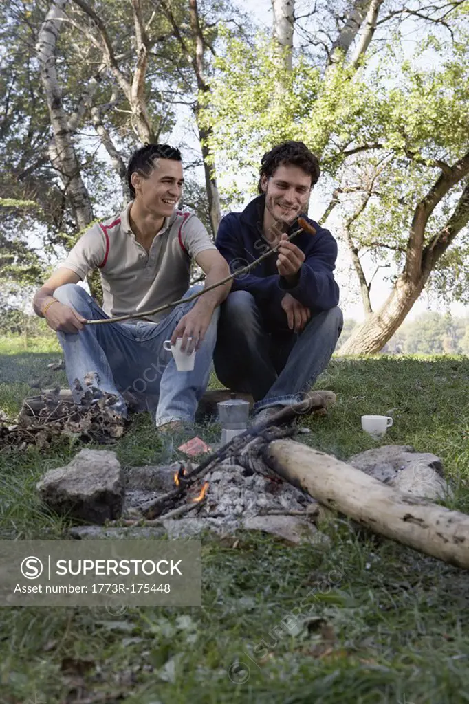 Two men cooking hot dogs over a fire pit smiling
